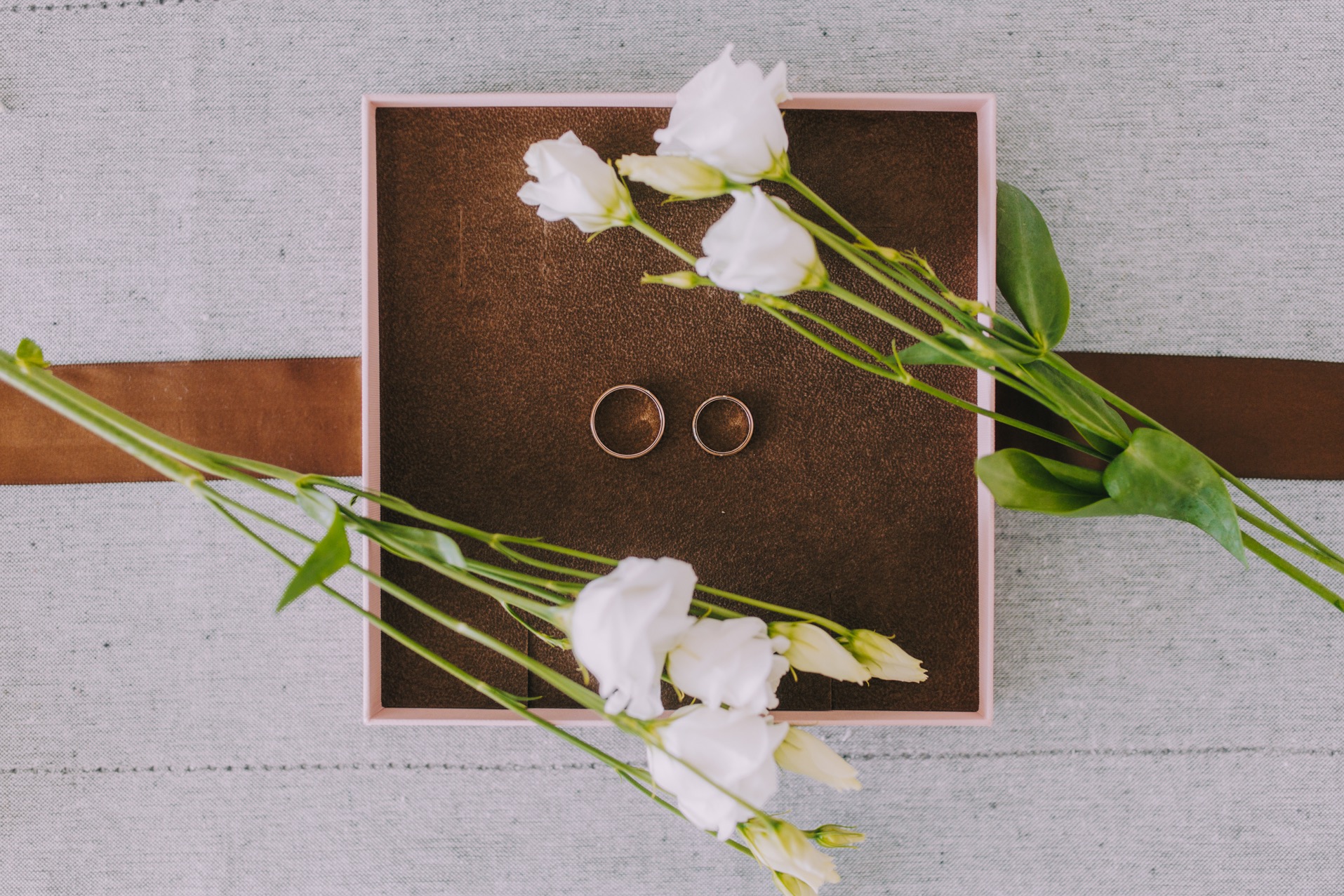 Dwa złote obrączki ślubne w brązowym pudełku z różową obwódką, otoczone białymi kwiatami na szarym tle z brązowym paskiem.