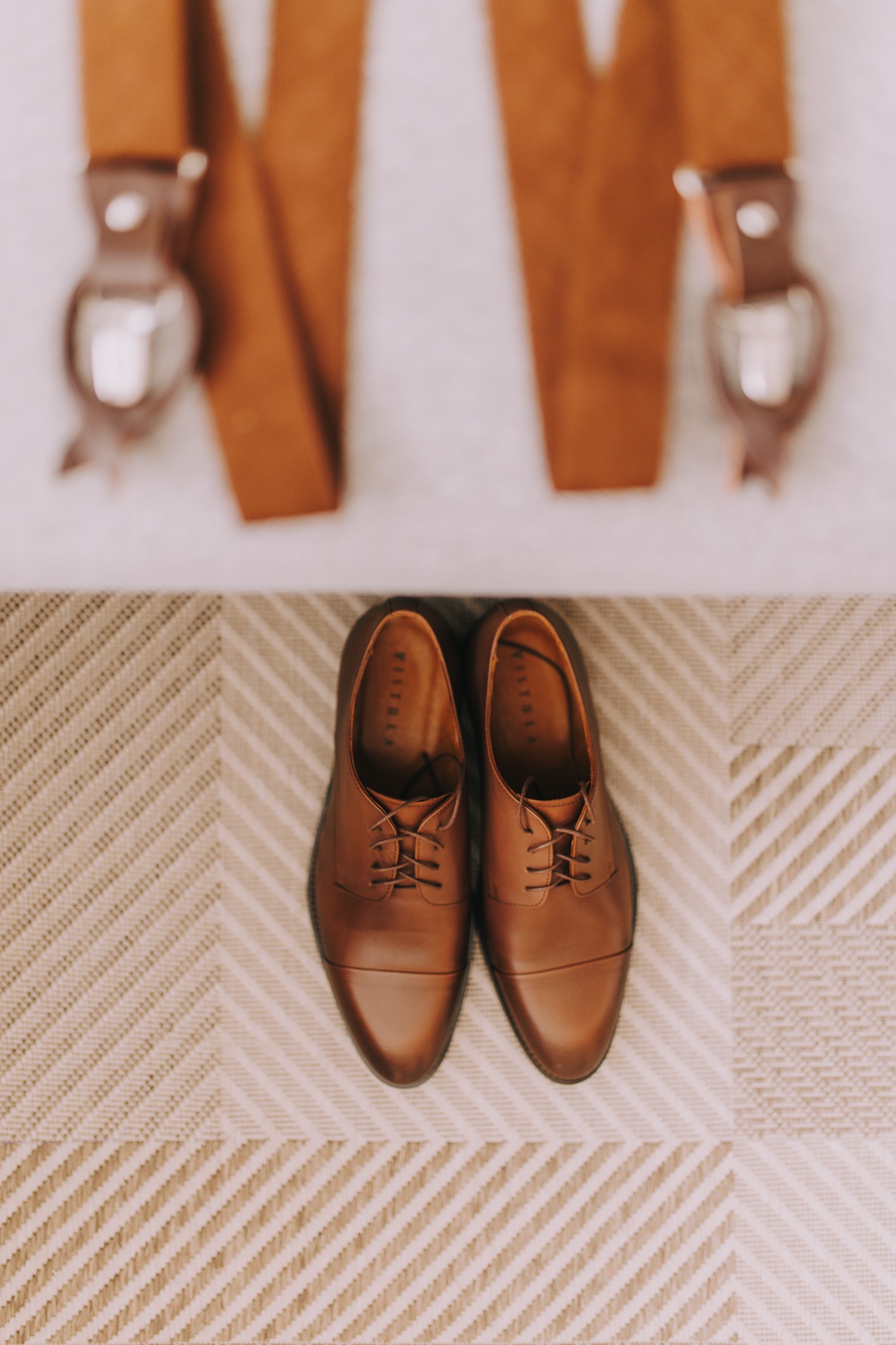 Brązowe, skórzane buty męskie z sznurowadłami ustawione na wzorzystym dywanie, nad nimi szelki z metalowymi klipsami, ujęcie z góry.