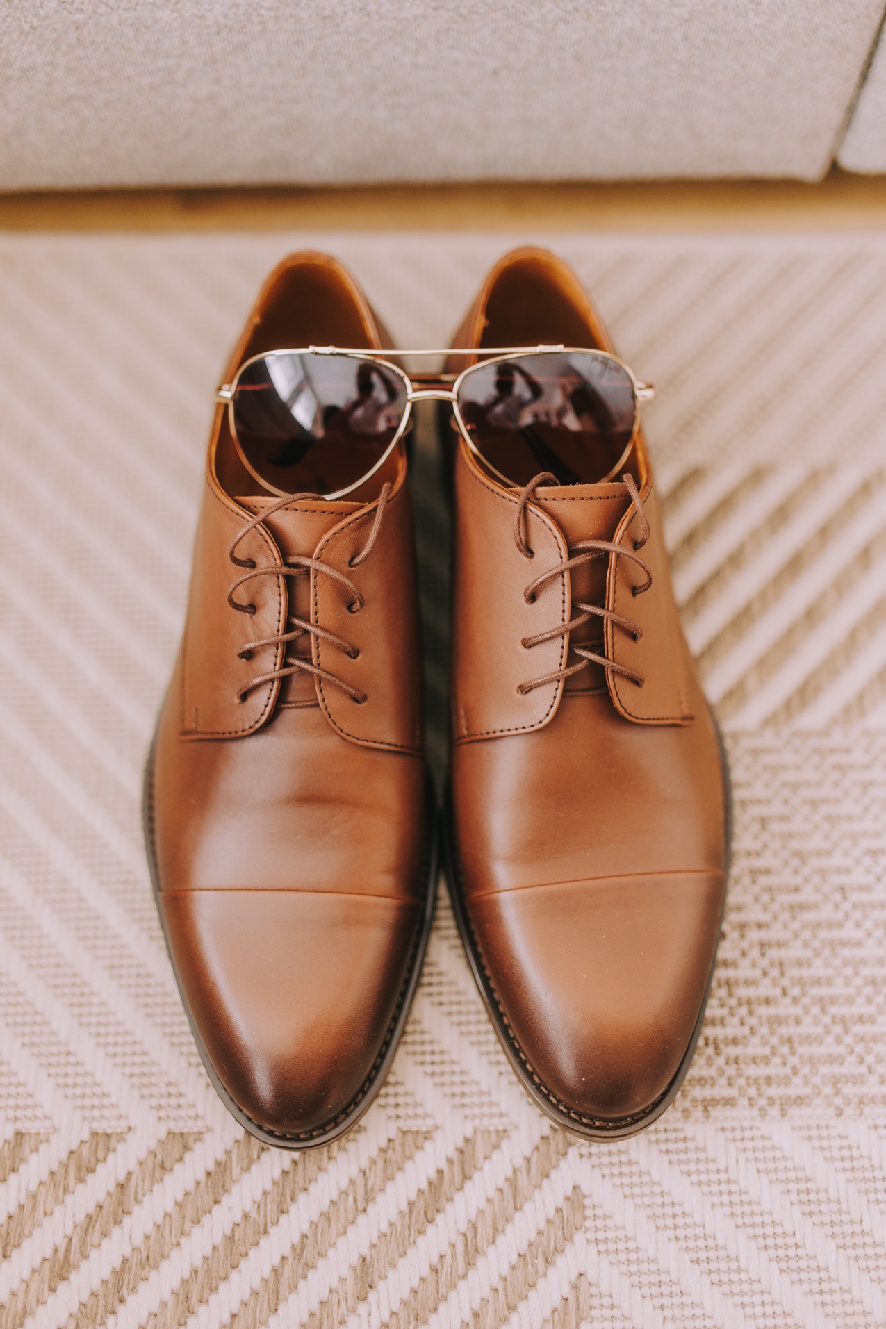 Brązowe, skórzane buty męskie ze sznurówkami, na których leżą złote okulary przeciwsłoneczne typu aviator, ułożone na beżowym dywanie w zygzakowaty wzór.