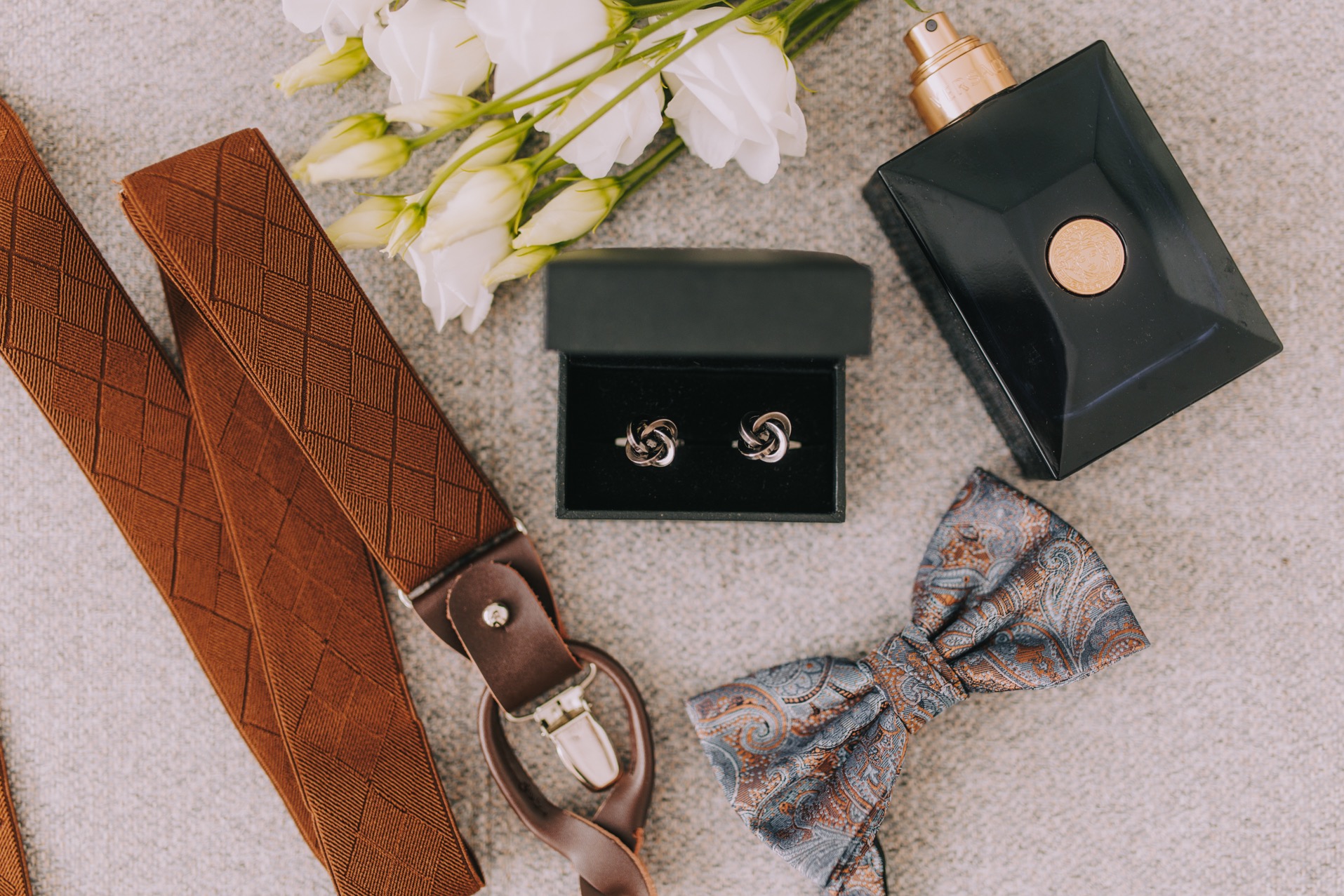 Męskie akcesoria ślubne: brązowe szelki z fakturą w romby, spinki do mankietów w czarnym pudełku, wzorzysta mucha, czarny flakon perfum i białe kwiaty na teksturowanym tle.
