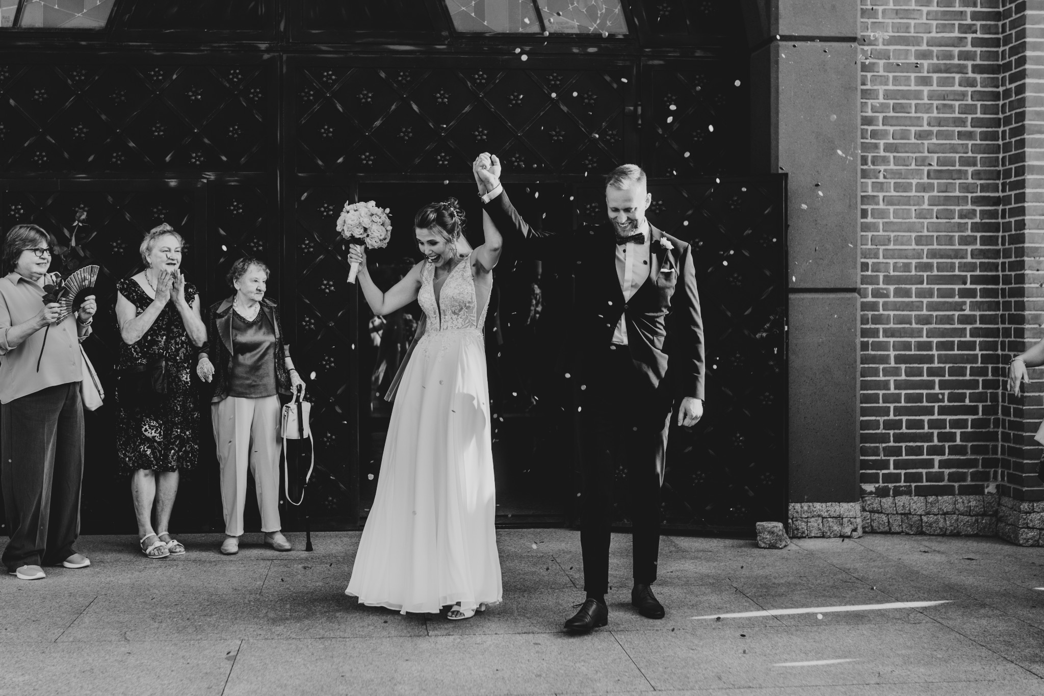 Czarno-białe zdjęcie nowożeńców wychodzących z budynku, obsypywanych konfetti, z uśmiechem na twarzy i uniesionymi rękami, w tle widoczne starsze kobiety.
