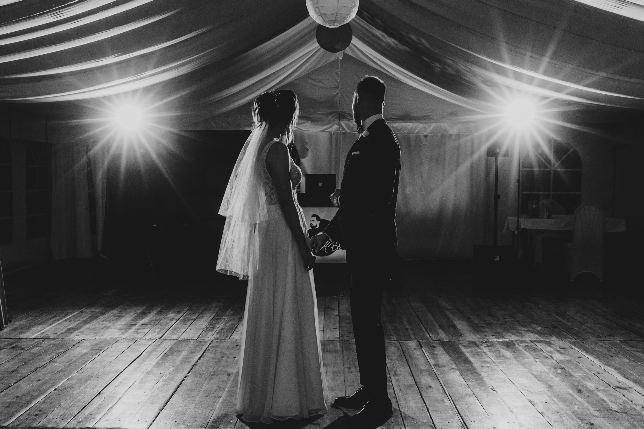 Silhouetted bride and groom holding hands under a tent with bright lighting, black and white photo on a wooden floor.