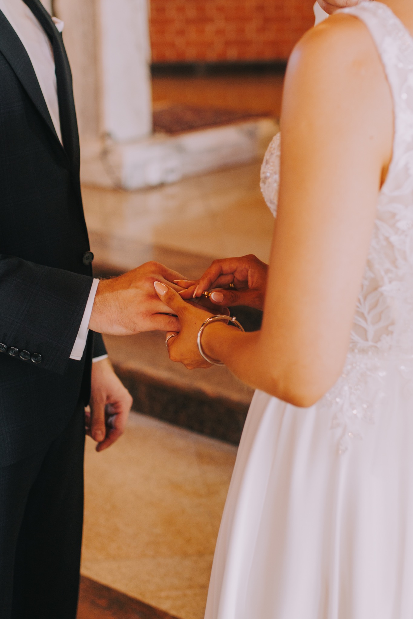 Panna młoda zakłada obrączkę panu młodemu podczas ceremonii ślubnej, widoczne dłonie, mankiet garnituru i fragment sukni ślubnej z koronkowymi aplikacjami.
