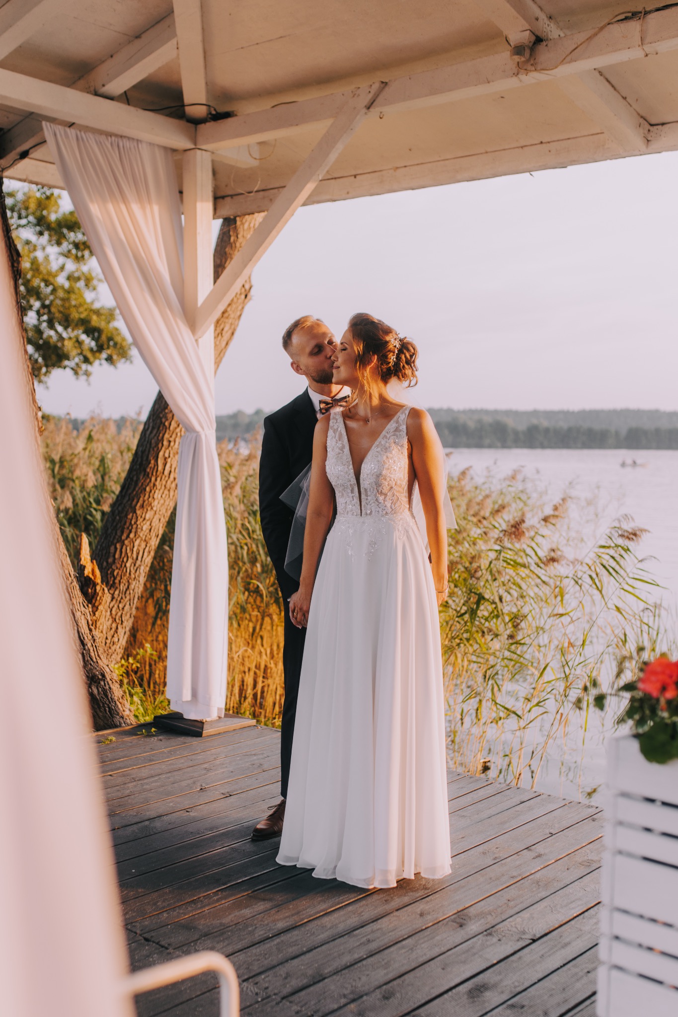 Panna młoda w białej sukni z koronkową górą i pan młody w czarnym garniturze całujący ją w skroń na drewnianym pomoście z widokiem na jezioro i trzciny w tle, w altanie z białymi zasłonami.