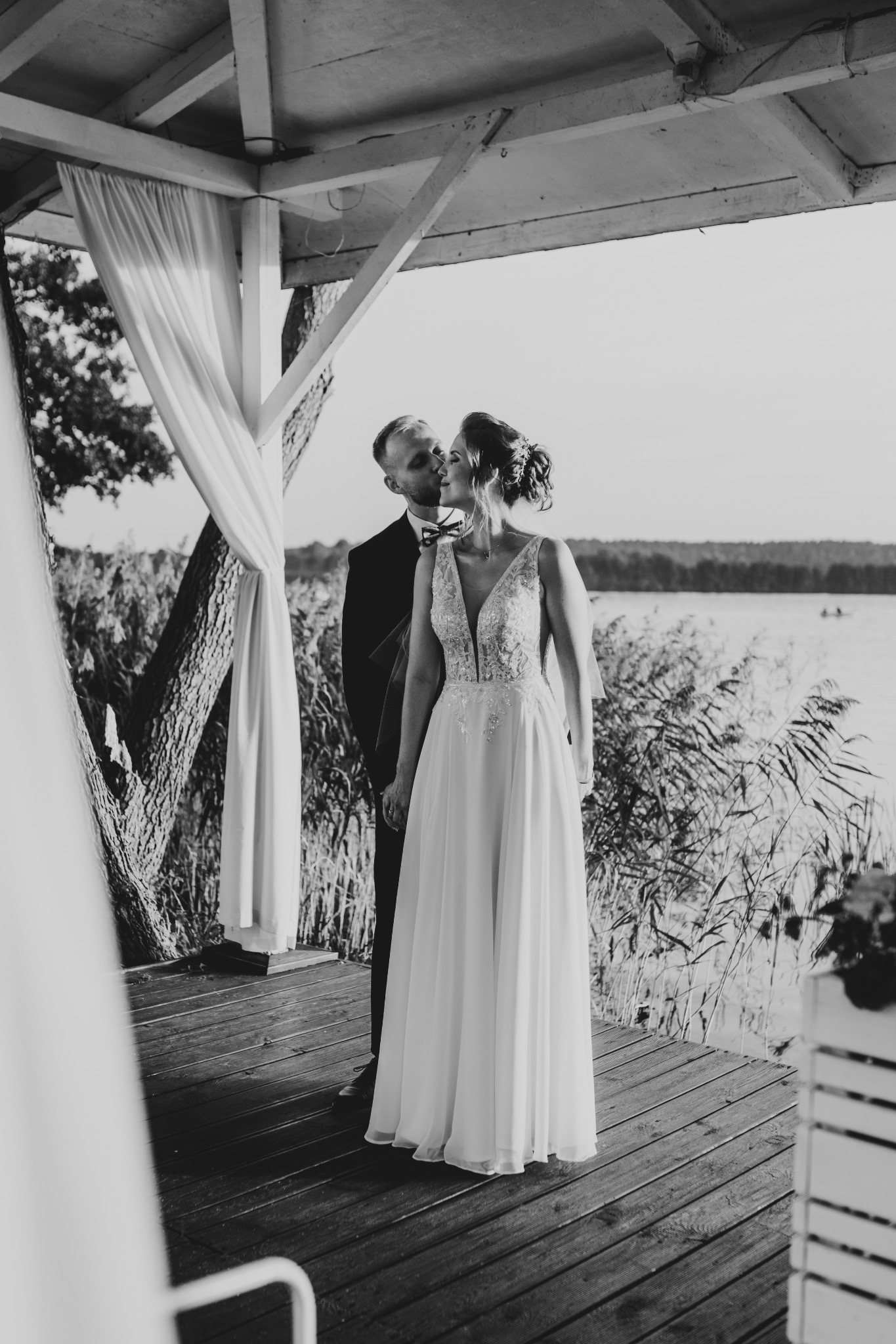 Czarno-białe zdjęcie pary młodej na drewnianym pomoście z widokiem na jezioro, panna młoda w sukni z koronkową górą, pan młody w czarnym garniturze z muszką, w tle trzciny i drzewa, dekoracja...