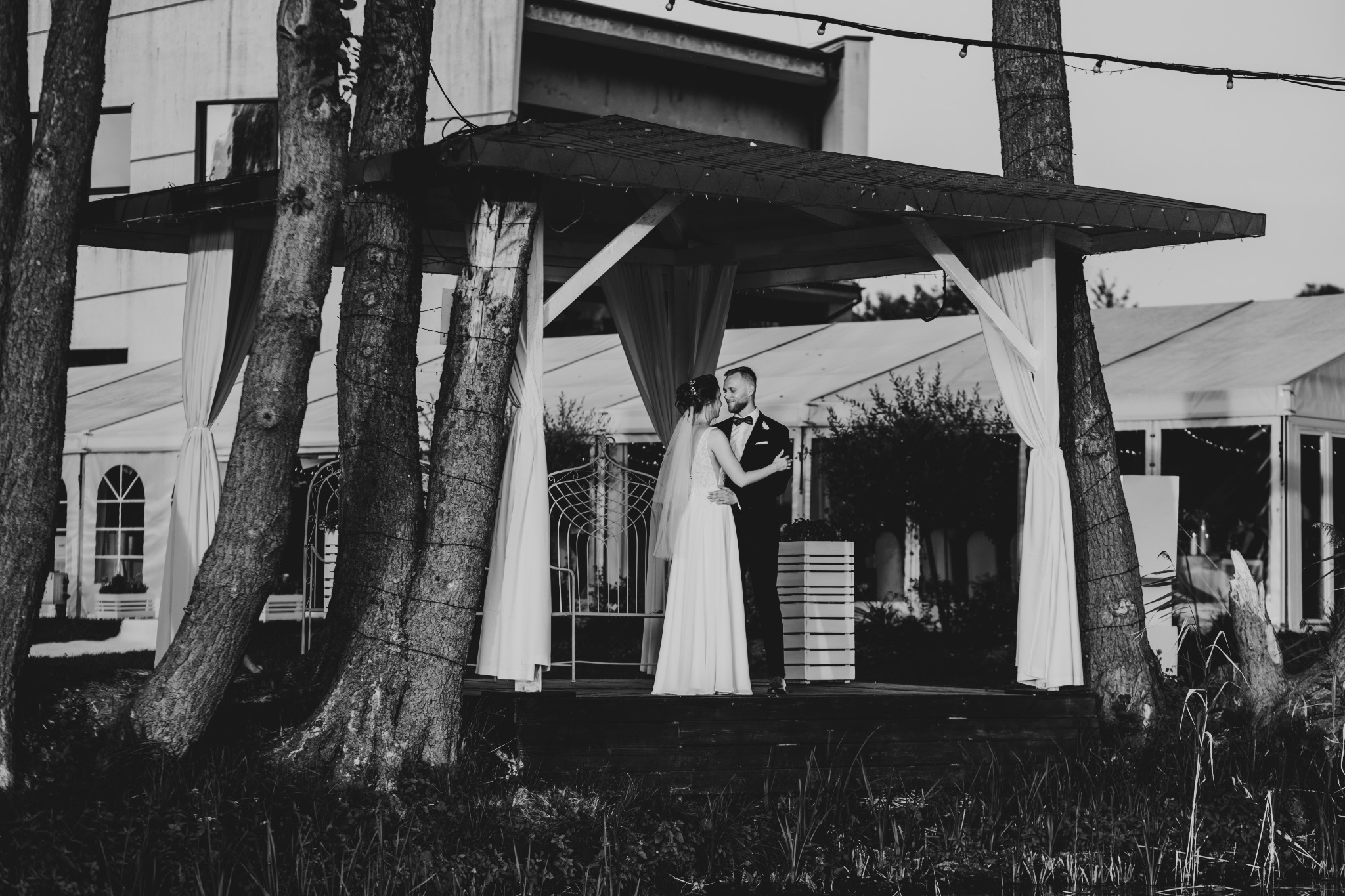 Czarno-białe zdjęcie pary młodej w objęciach pod drewnianą altaną z białymi zasłonami, drzewa wkomponowane w konstrukcję, w tle budynek z oknami.