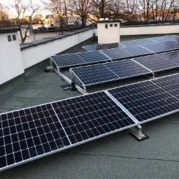 Panele fotowoltaiczne zamontowane na płaskim dachu budynku w Jaworniku, widoczne kominy wentylacyjne i drzewa w tle.