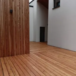 Nowoczesny taras z desek kompozytowych w odcieniach brązu, przylegający do białej ściany budynku z minimalistycznym oknem i widocznym fragmentem drewnianej elewacji. Widoczny fragment dachu...