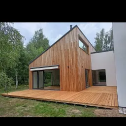 Nowoczesny dom jednorodzinny z drewnianą elewacją i dużym tarasem, otoczony zielenią, widok z zewnątrz.