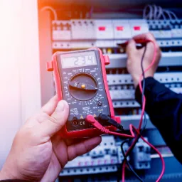 Elektryk mierzy napięcie w rozdzielnicy elektrycznej za pomocą multimetru z czerwonym korpusem, wskazującym wartość 234 na wyświetlaczu LCD.