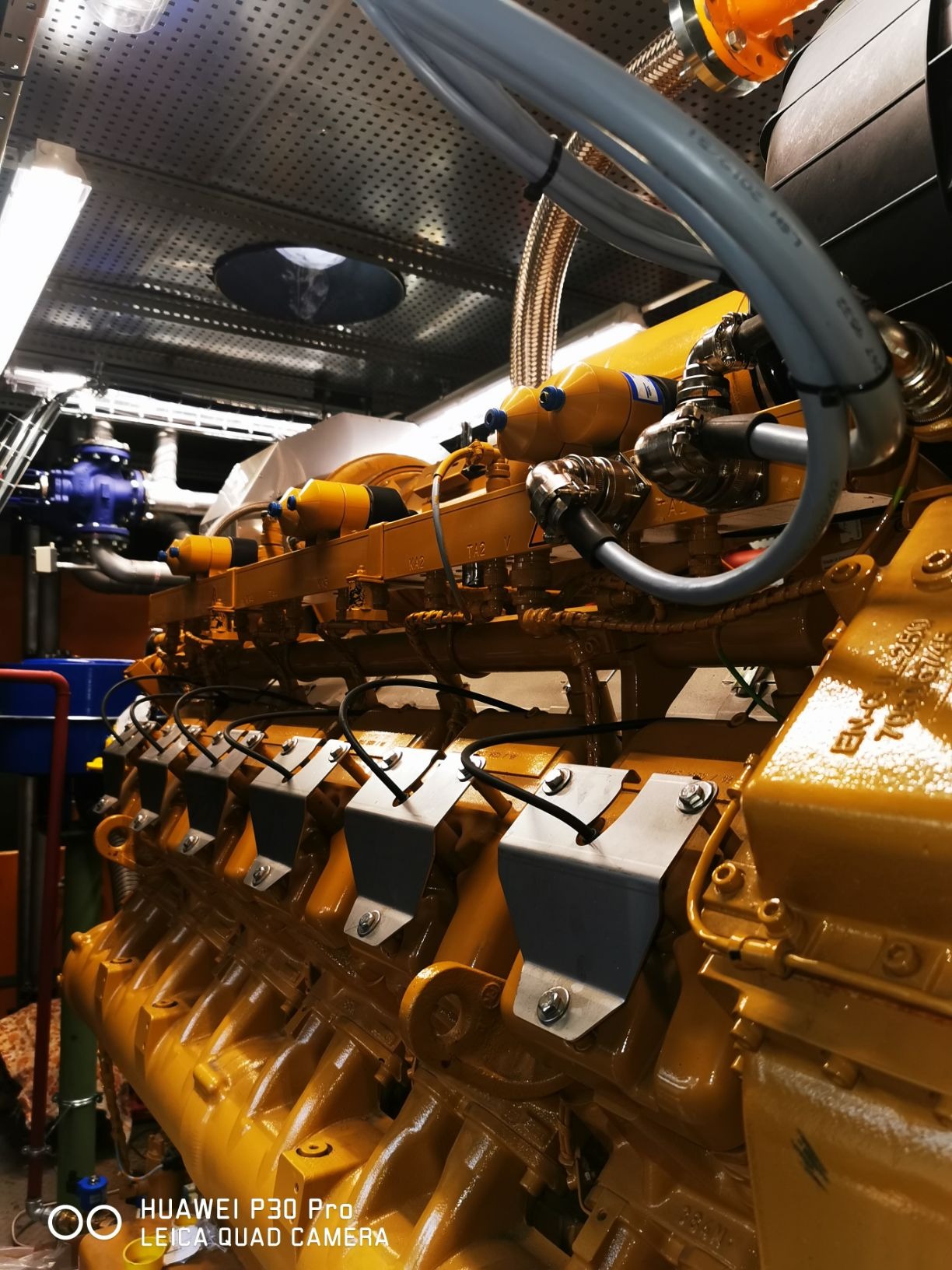 Żółty silnik przemysłowy z metalowymi osłonami, widoczne przewody, rury i elementy instalacji w pomieszczeniu technicznym.