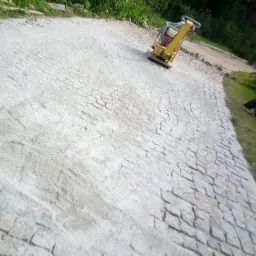 Żółta zagęszczarka wibrująca ubija świeżo ułożoną, jasnoszarą kostkę brukową na podjeździe, widok z boku, zieleń w tle.