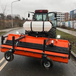 Instytut prac porządkowych Sp zoo - Pomarańczowa szczotka czyszcząca zamontowana na traktorze, gotowa do pracy na mokrej, asfaltowej drodze z widocznymi śladami opon i błota, w tle budynki mieszkalne i drzewa bez liści.