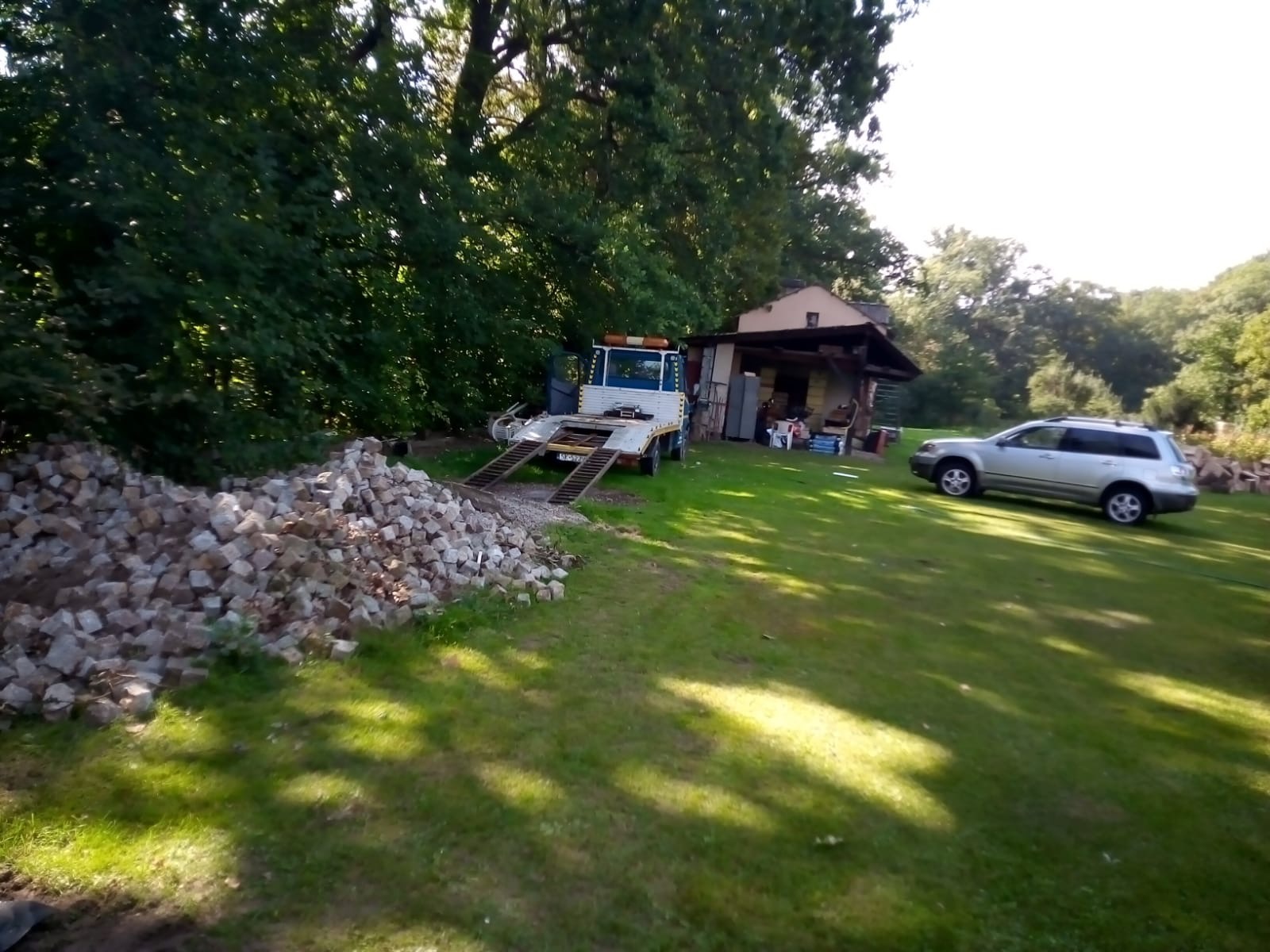 Samochód transportowy z najazdem zaparkowany na trawniku obok sterty kostki brukowej, w tle mały budynek gospodarczy i samochód osobowy.