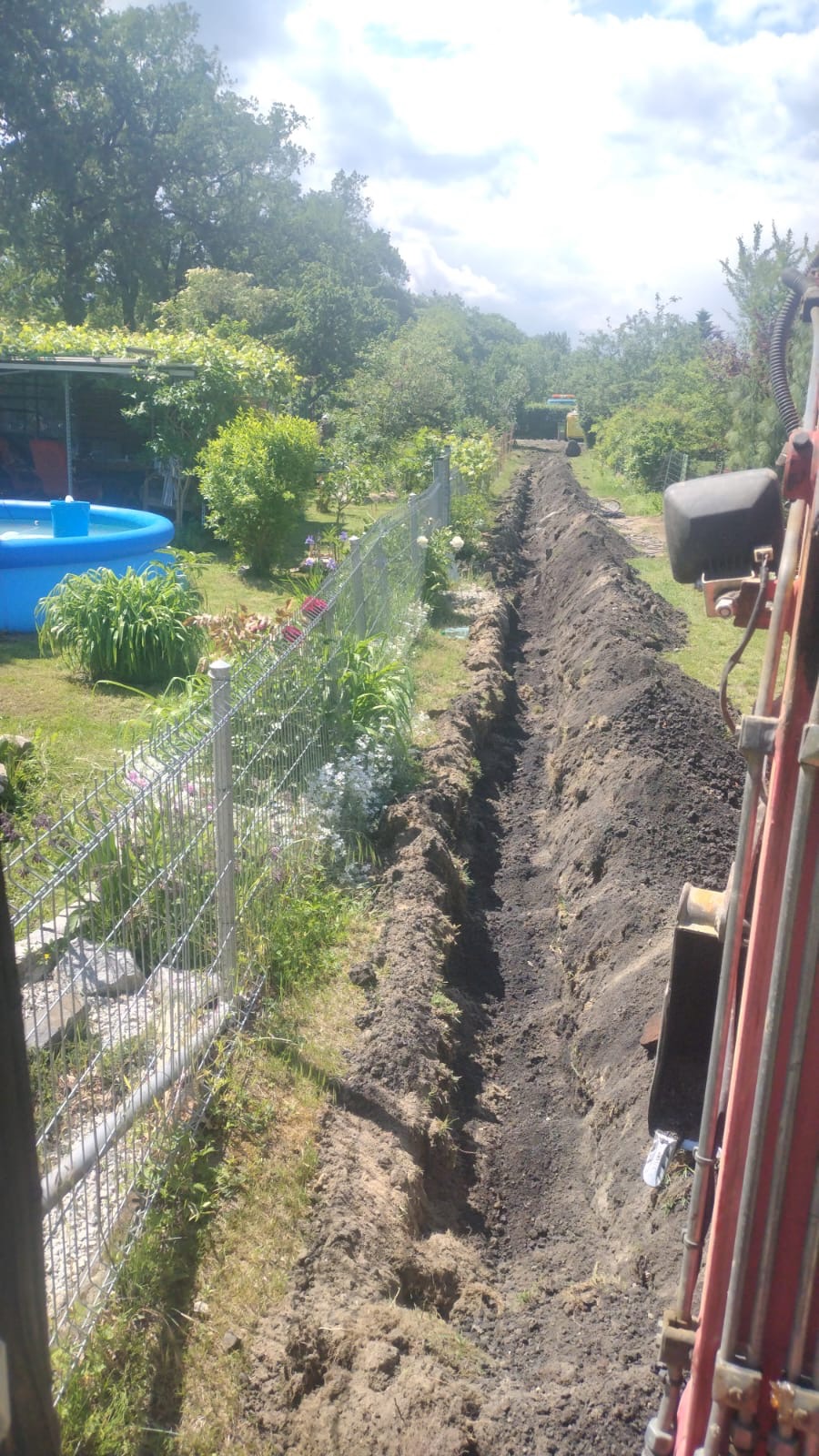 Wąski wykop wzdłuż ogrodzenia panelowego w ogrodzie, z widoczną koparką i basenem w tle.