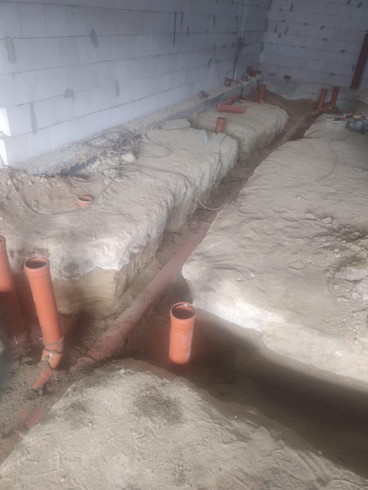 Wykopane rowy w ziemi z ułożonymi rurami kanalizacyjnymi z tworzywa sztucznego wewnątrz budynku z białych bloczków.