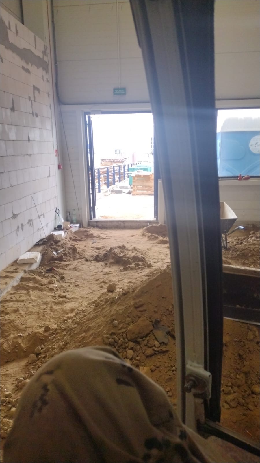 Wnętrze hali w trakcie przygotowania pod wylewkę, widoczny piasek, fragment ściany z bloczków betonowych oraz otwarte drzwi z widokiem na plac budowy.