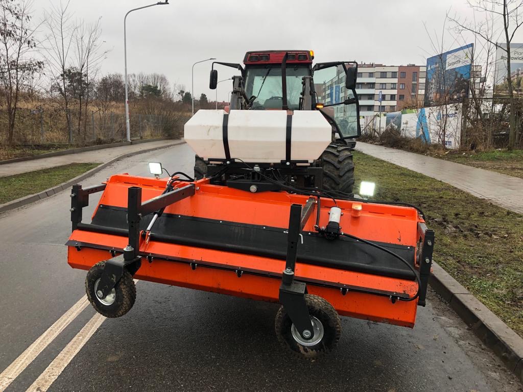 Pomarańczowa szczotka czyszcząca zamontowana na traktorze, gotowa do pracy na mokrej, asfaltowej drodze z widocznymi śladami opon i błota, w tle budynki mieszkalne i drzewa bez liści.