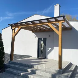 Drewniana konstrukcja z przezroczystym zadaszeniem nad wejściem do domu, schody wyłożone kostką brukową, elewacja budynku w jasnym kolorze, błękitne niebo.