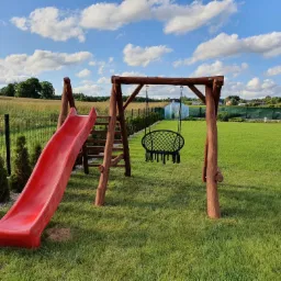 Plac zabaw z drewnianą konstrukcją huśtawki z czarnym siedziskiem typu bocianie gniazdo i czerwoną zjeżdżalnią na trawniku, w tle pole i błękitne niebo z chmurami.
