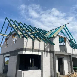 Budowa domu jednorodzinnego z bloczków, na etapie montażu drewnianej, pomalowanej na zielono konstrukcji dachu, widok z dołu na tle błękitnego nieba z chmurami.