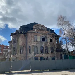 Stary, częściowo odrestaurowany budynek z widocznymi śladami zniszczeń, zabezpieczony szarym płotem z blachy falistej, z nowym, ciemnym dachem i odsłoniętą cegłą na elewacji.