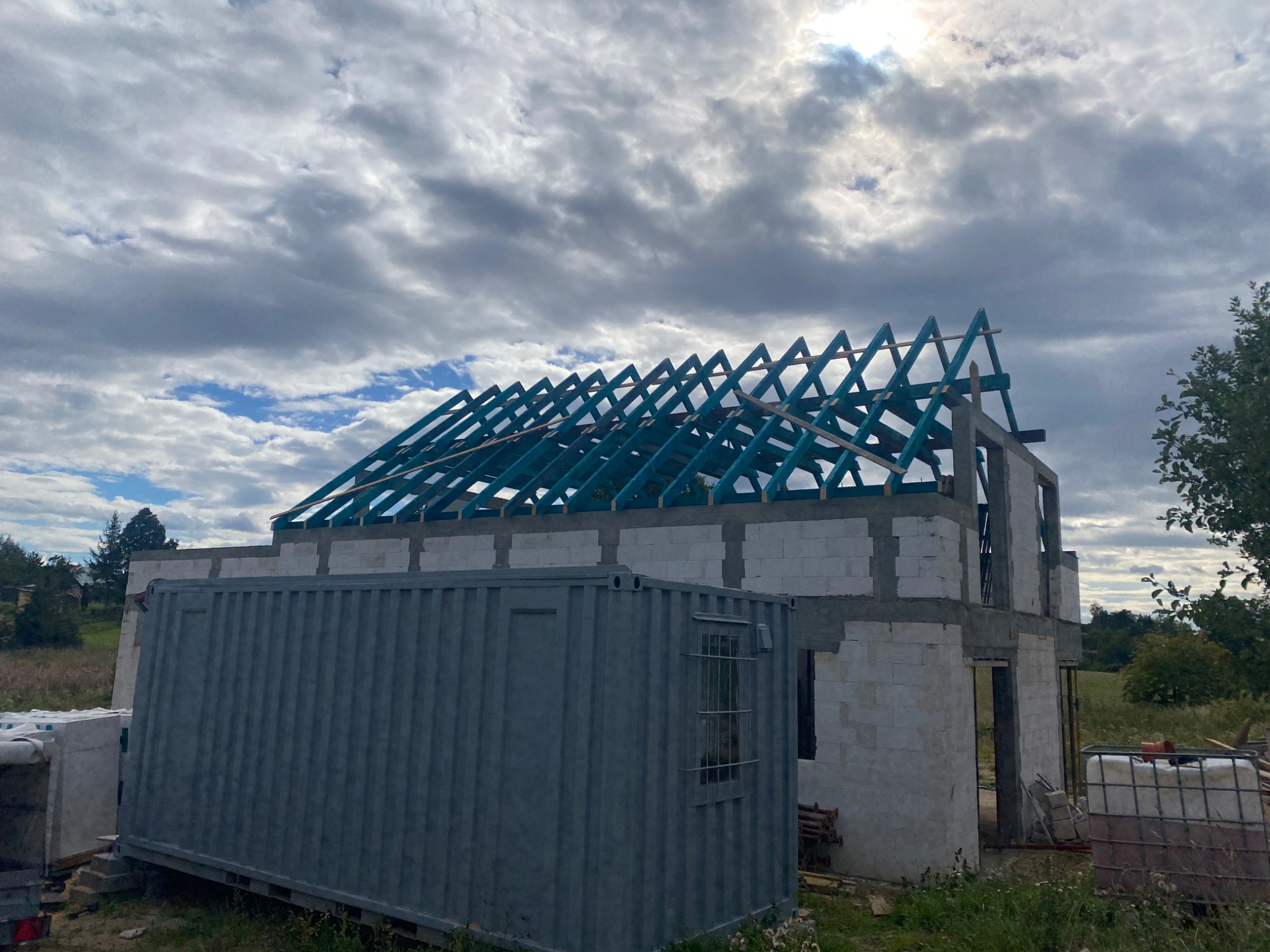 Budowa domu w trakcie wznoszenia, widoczna konstrukcja ścian z betonu komórkowego i częściowo zmontowana, pomalowana na niebiesko więźba dachowa; w tle pochmurne niebo i fragment krajobrazu,...