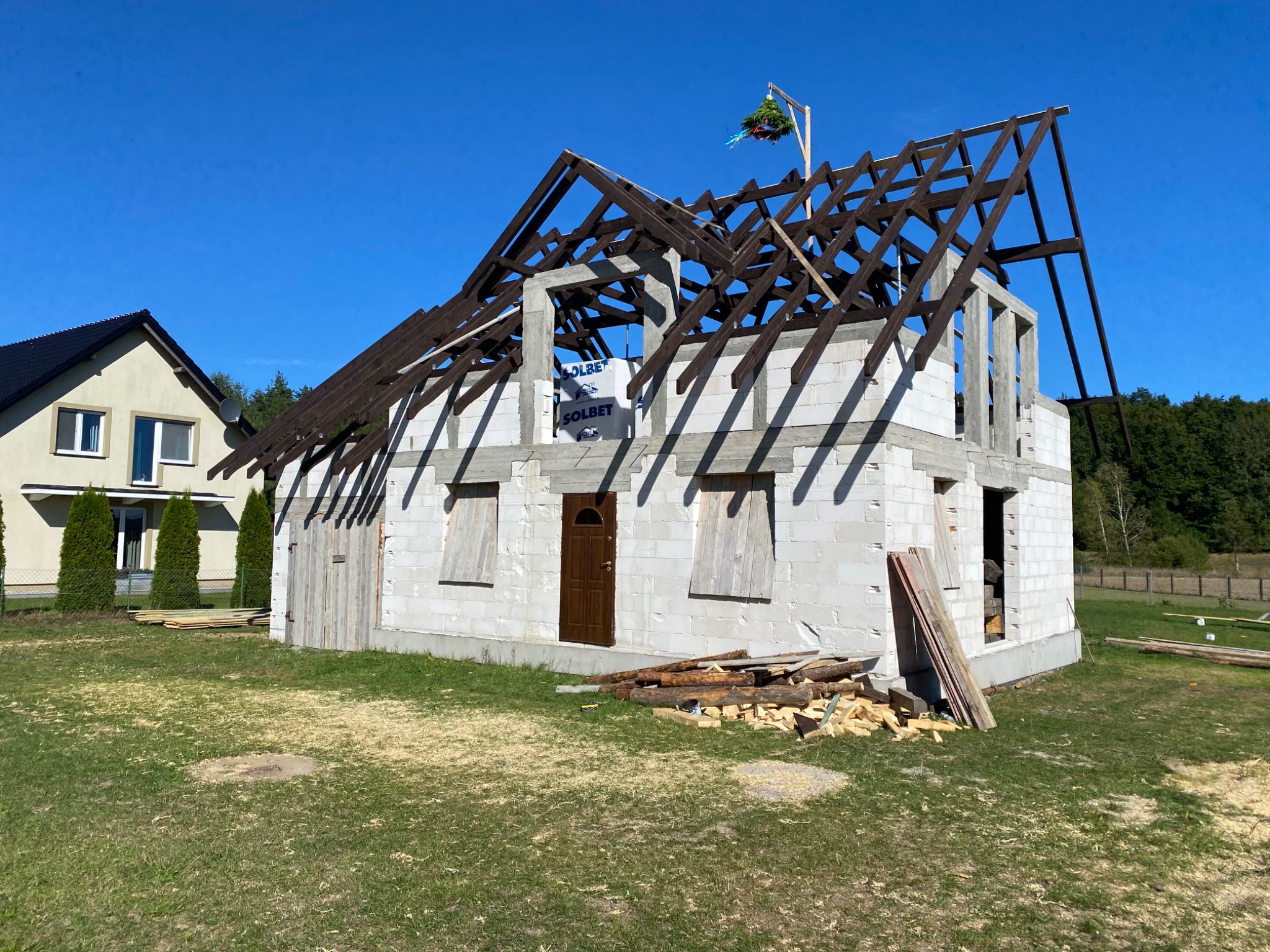 Wznoszenie domu z pustaków: widoczna surowa konstrukcja ścian, brązowe elementy więźby dachowej, wieniec na szczycie dachu, obok drugi dom i stos drewna na trawie.