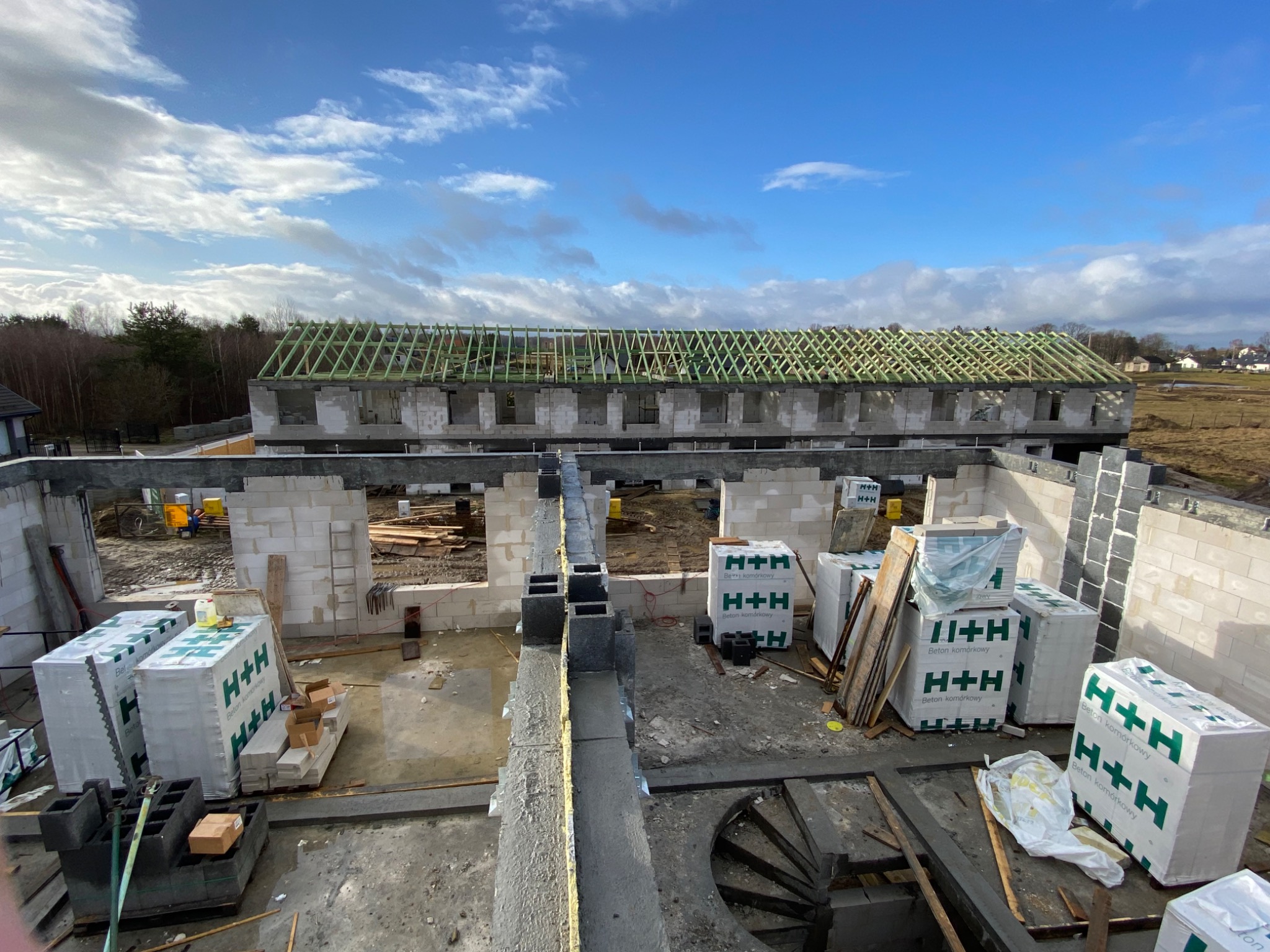 Wznoszenie ścian z bloczków betonowych komórkowych H+H na budowie domu jednorodzinnego, widoczna konstrukcja drewniana dachu w trakcie montażu, błękitne niebo z chmurami w tle.