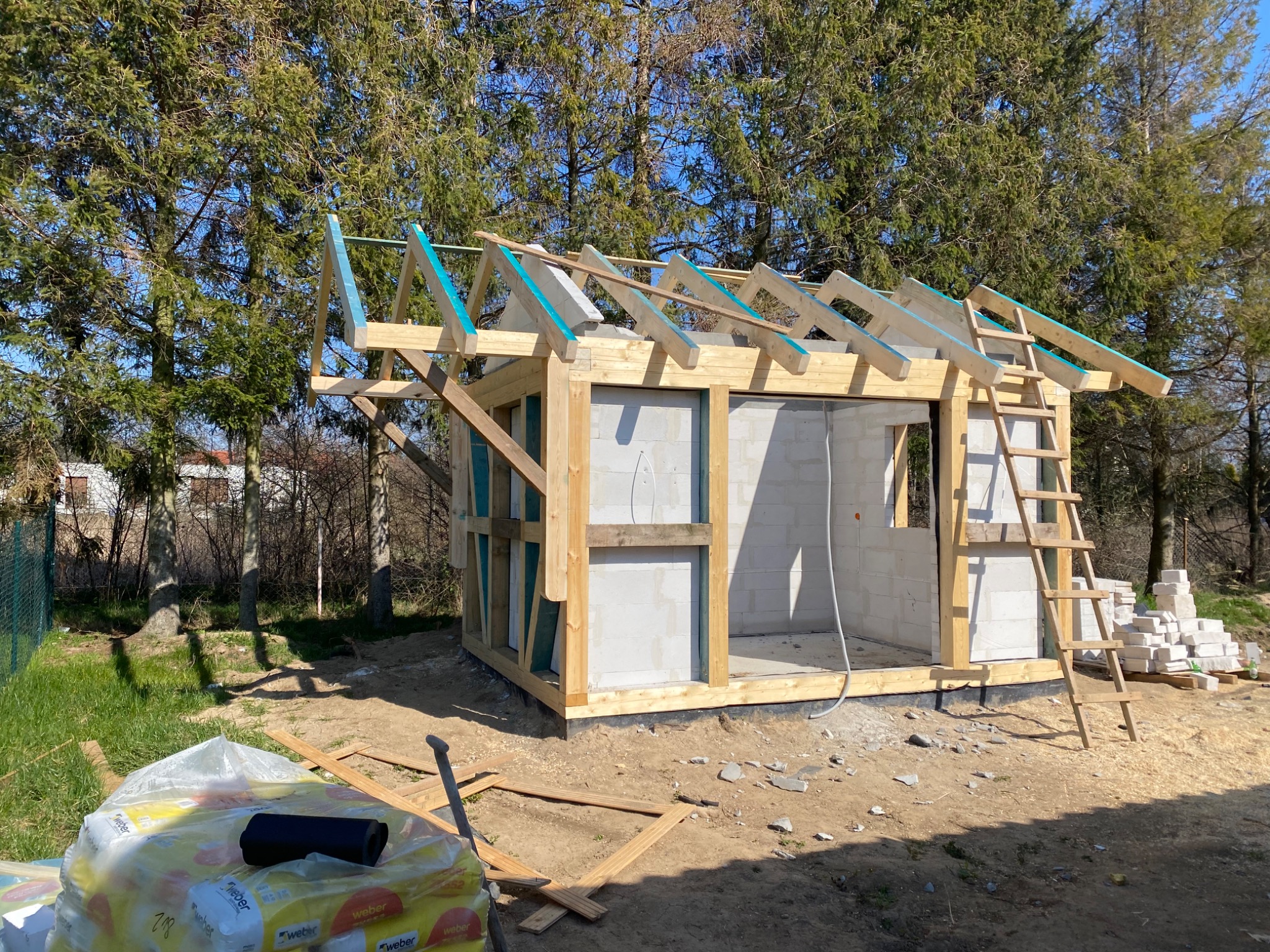 Surowy szkielet małego budynku z bloczków i drewna, z widoczną konstrukcją dachu w trakcie montażu, drabina oparta o ścianę, paczki zaprawy w tle, otoczenie drzewa i trawa.