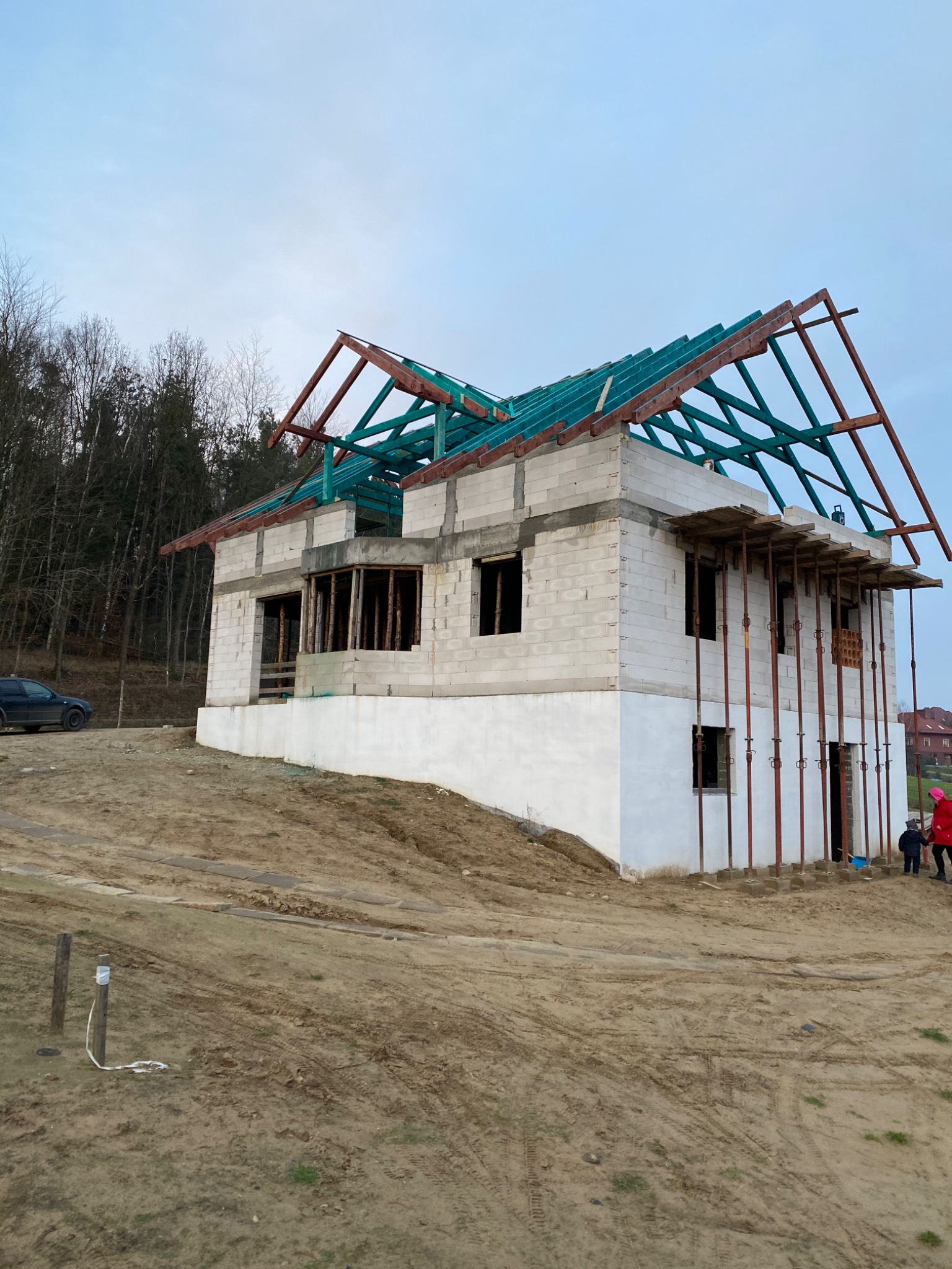 Budowa domu jednorodzinnego w stanie surowym otwartym: widoczna konstrukcja ścian z bloczków, częściowo otynkowana podmurówka, drewniana więźba dachowa z zielonymi elementami impregnacji, stalowe...