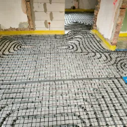 Instalacja ogrzewania podłogowego: szare rury ułożone w pętle na czarnej siatce, żółta taśma brzegowa przy ścianach w surowym stanie.