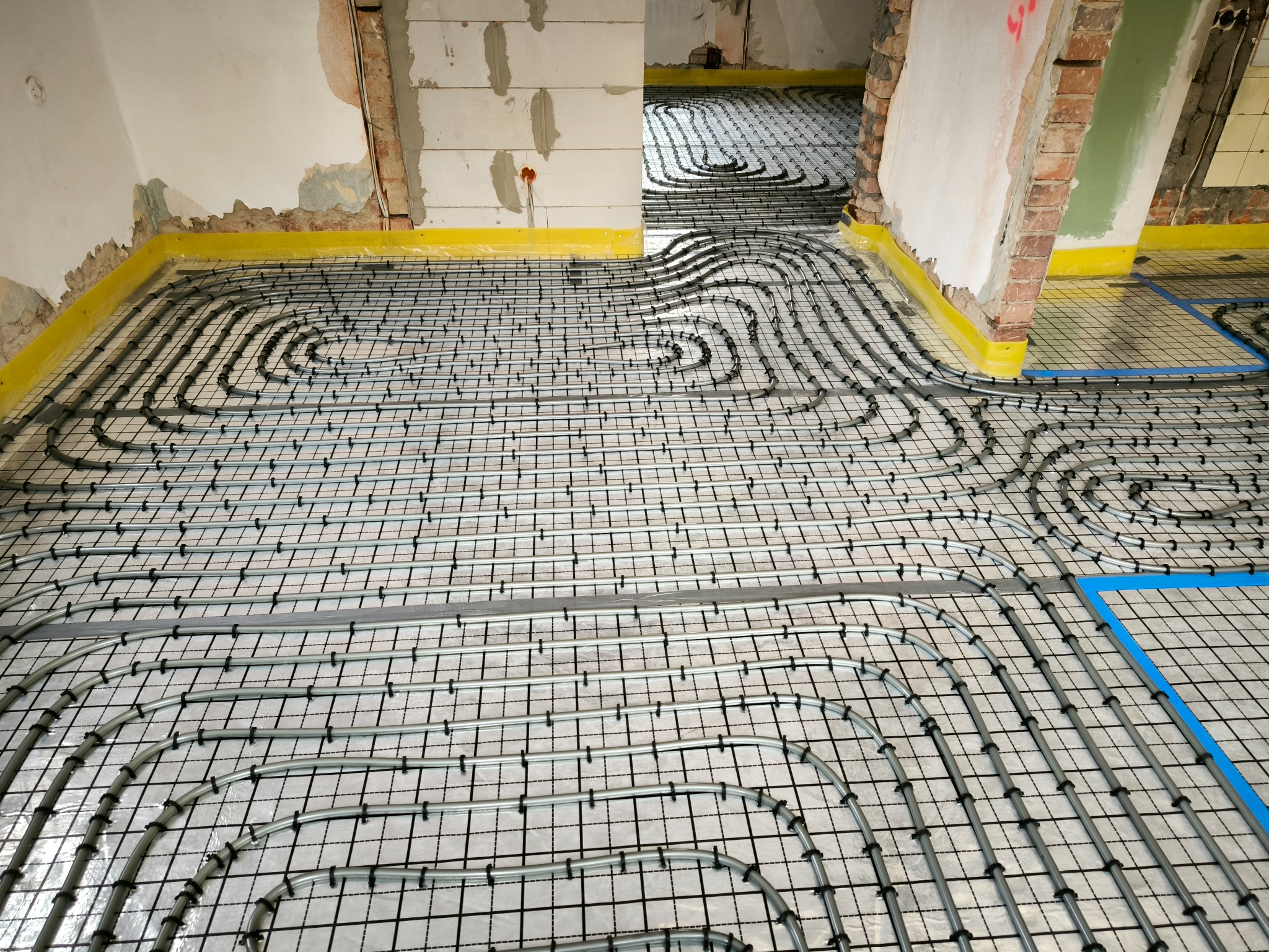 Instalacja ogrzewania podłogowego: szare rury ułożone w pętle na czarnej siatce, żółta taśma brzegowa przy ścianach w surowym stanie.