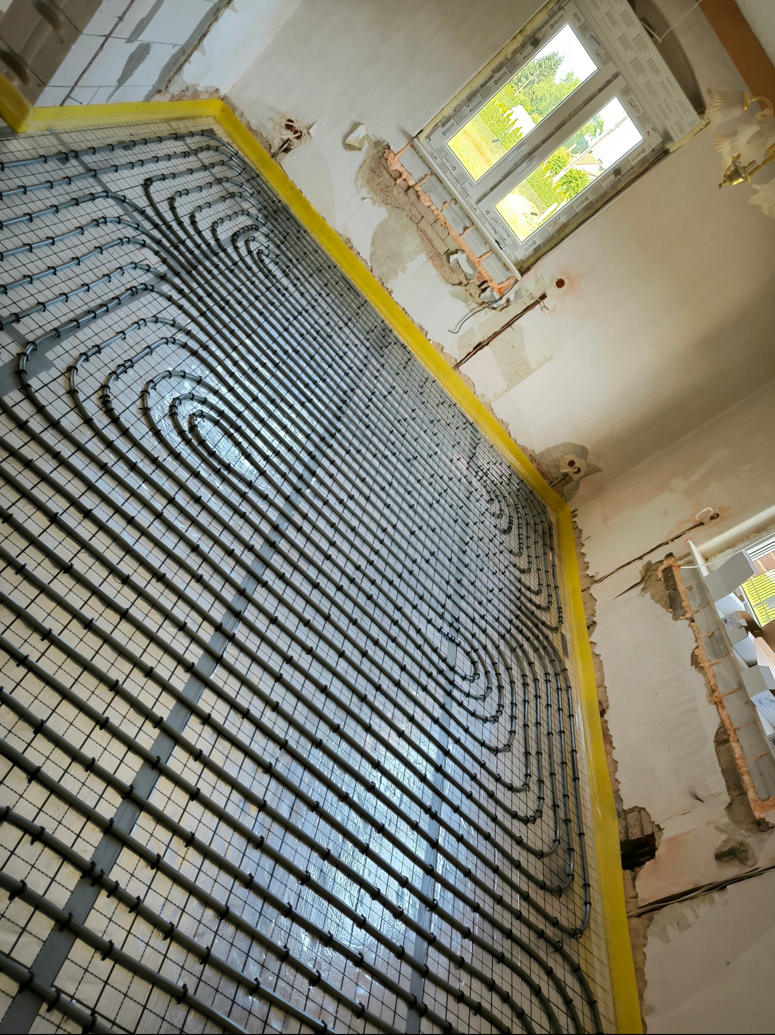 Widok pod kątem na instalację ogrzewania podłogowego w trakcie montażu, rury ułożone w pętle na izolacji, przy ścianie żółta taśma brzegowa, w tle okno i surowy stan wykończenia wnętrza.