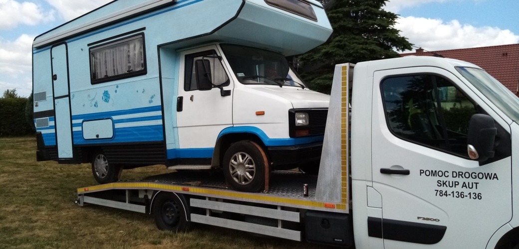 Biały kamper z niebieskimi akcentami, z delikatnymi kwiatowymi wzorami, transportowany na platformie pomocy drogowej z napisem 'Pomoc Drogowa Skup Aut'.