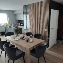 Jadalnia z drewnianym stołem, aksamitnymi krzesłami i dekoracyjną ścianą z drewnianych listew, widok na kuchnię w tle.