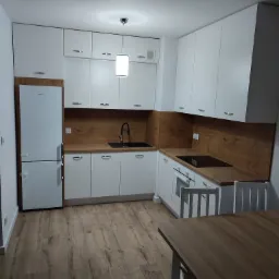 Nowoczesna kuchnia z białymi szafkami i drewnianym blatem, czarną armaturą i wiszącą lampą, uzupełniona o drewniany stół z białymi krzesłami na parkiecie.