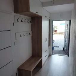 Przedpokój w Rumi z zabudową na wymiar: białe szafy z drewnianymi akcentami, siedzisko z wieszakami i widok na zewnątrz przez otwarte drzwi.