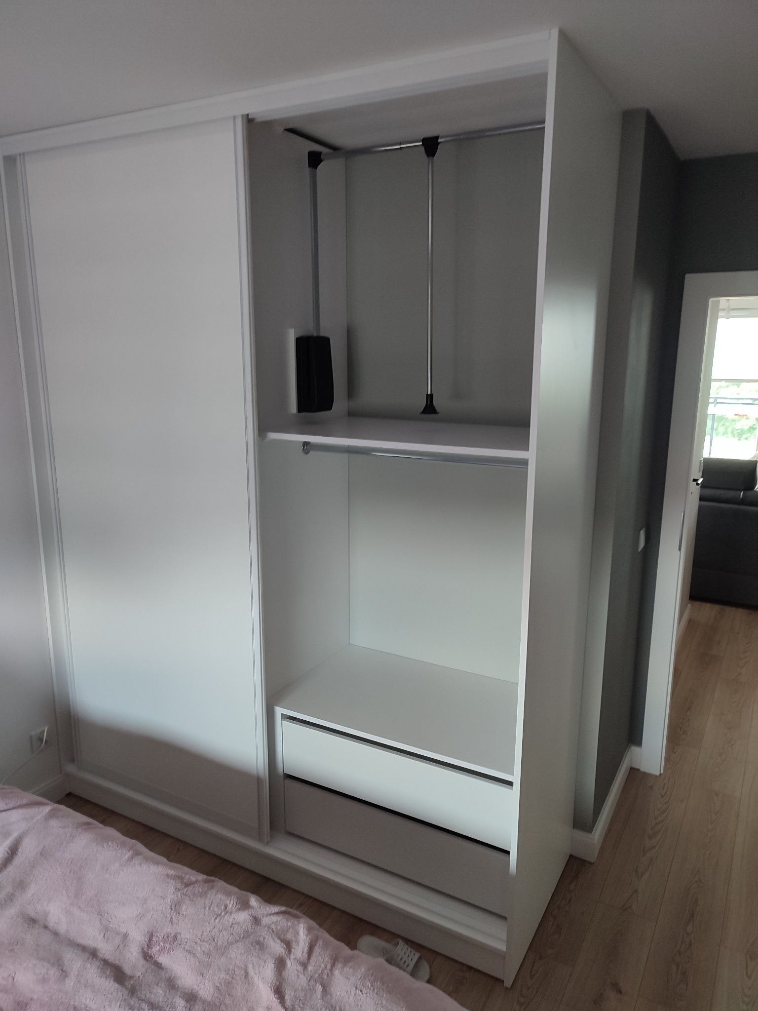 Biała szafa przesuwna z otwartą częścią, widoczne półki, szuflady i drążki na ubrania, wnętrze w odcieniach szarości, fragment sypialni z łóżkiem i drewnianą podłogą.