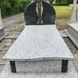 Jasny granitowy nagrobek z ciemnymi, łezkowatymi elementami bocznymi i krzyżem z wizerunkiem Chrystusa, otoczony innymi nagrobkami na placu kamieniarskim.