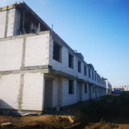 Wznoszenie szeregu domów z bloczków z betonu komórkowego, widoczne szalunki i zbrojenia w górnej części, teren budowy z ziemią i roślinnością w tle.