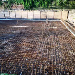 Siatka zbrojeniowa fundamentu z wystającymi prętami pionowymi, drewniane szalunki na obrzeżach wykopu, zabezpieczone deskami skarpy.