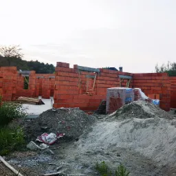 Wznoszone ściany domu z czerwonej cegły Leier, widoczne palety z cegłami, hałdy piasku i gruzu na placu budowy.