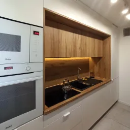 Nowoczesna kuchnia z białymi szafkami, drewnianym blatem i zabudową, czarnym zlewozmywakiem i płytą indukcyjną, oświetleniem LED pod szafkami wiszącymi i srebrnym czajnikiem.