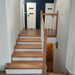 Nowe drewniane schody z jasnego drewna z białymi podstopnicami i drewnianą balustradą, prowadzące na górę, widoczne drzwi w tle.