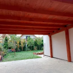 Zadaszenie tarasu z bejcowanego drewna, widok od spodu na drewnianą konstrukcję, w tle ogród z zielonym trawnikiem i budynkiem mieszkalnym.