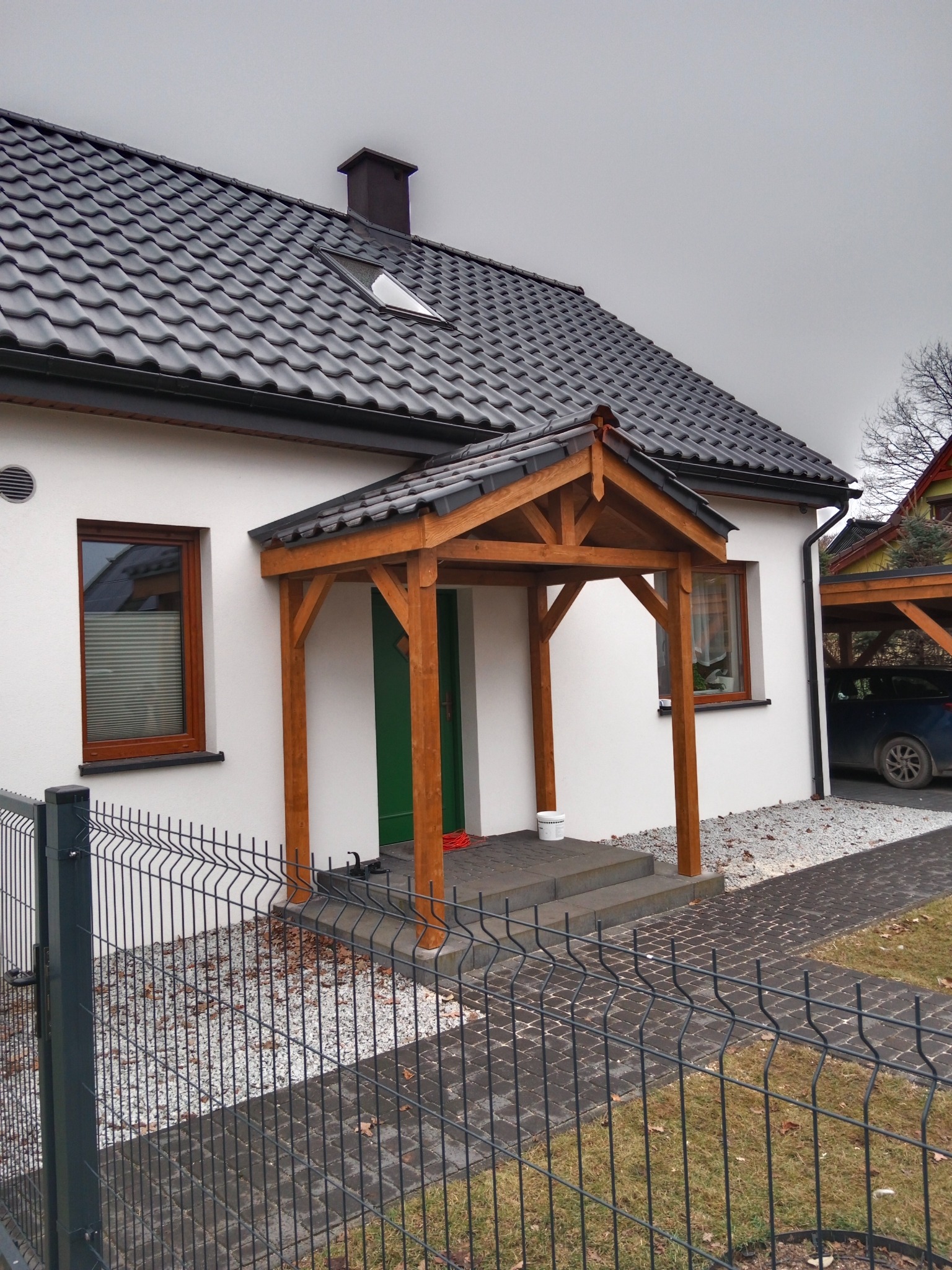 Biały dom z ciemną dachówką i drewnianym zadaszeniem nad wejściem, widoczne schody z szarej kostki, metalowe ogrodzenie i fragment podjazdu.