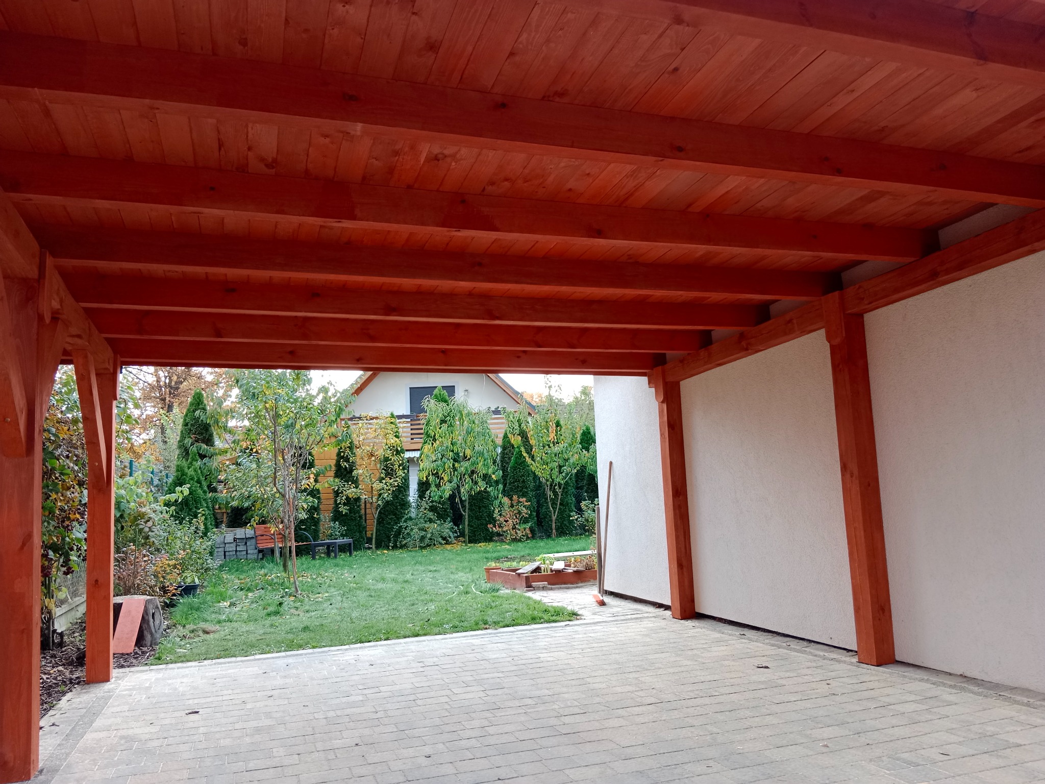 Zadaszenie tarasu z bejcowanego drewna, widok od spodu na drewnianą konstrukcję, w tle ogród z zielonym trawnikiem i budynkiem mieszkalnym.