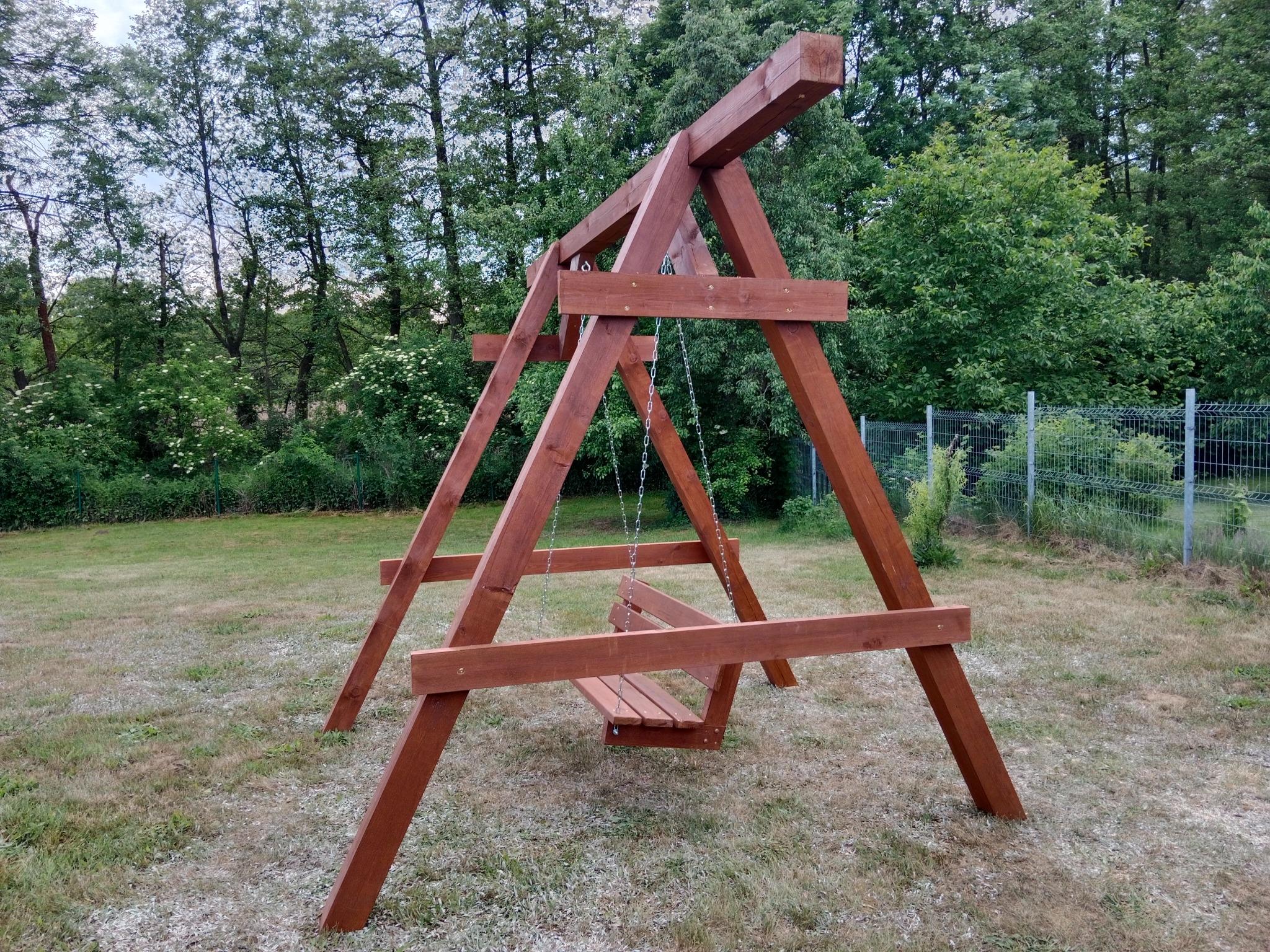 Drewniana huśtawka ogrodowa z bejcowanego drewna, zawieszona na metalowych łańcuchach, ustawiona na trawniku w ogrodzie.