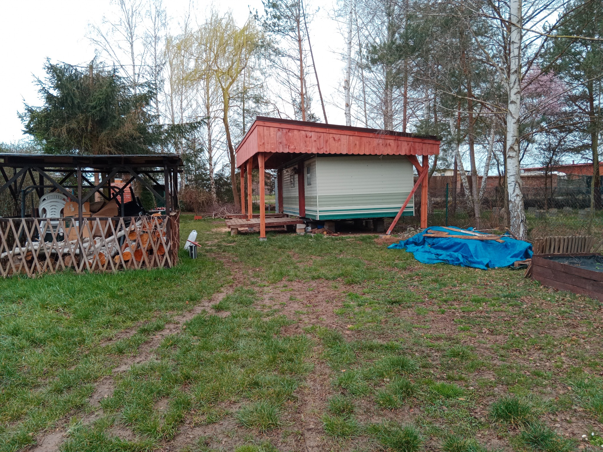 Niewielki, parterowy domek letniskowy z drewnianą konstrukcją dachu i werandy, widoczny z zewnątrz na działce z drzewami i stertą drewna opałowego.